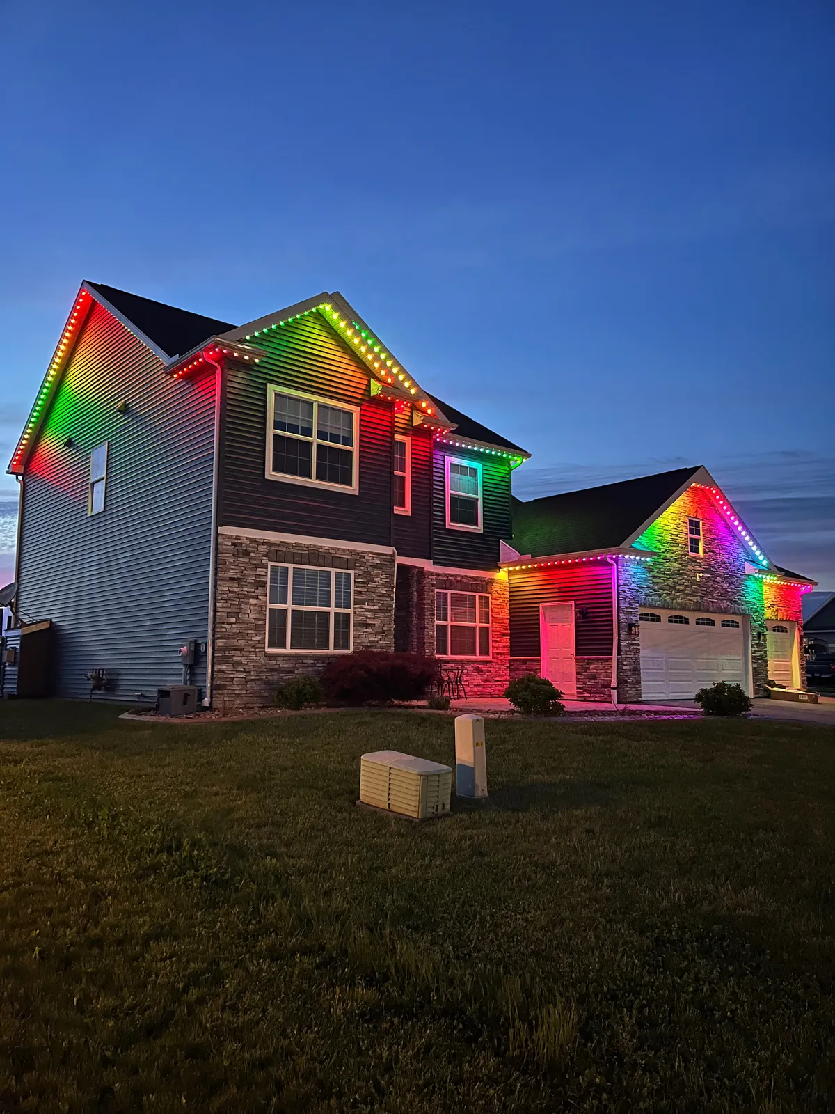 Two-story home with stone-front garage at dusk, permanent lights along every roofline glowing in a rainbow of colors
