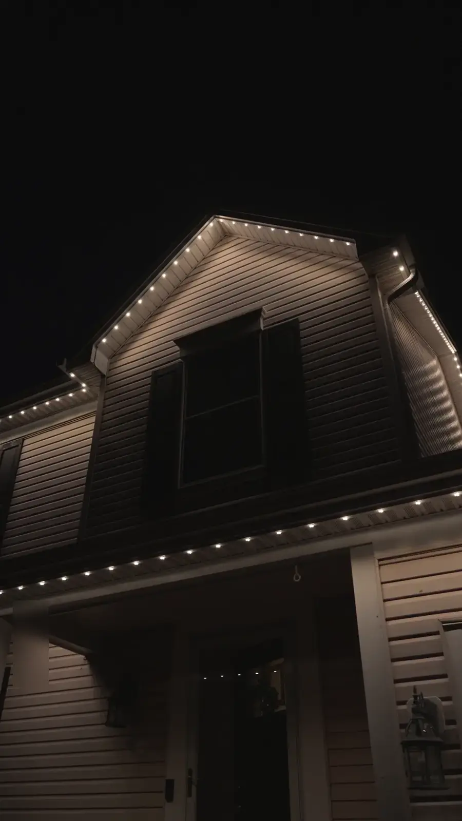The same house after dark with warm white permanent lights tracing the gable and porch roof