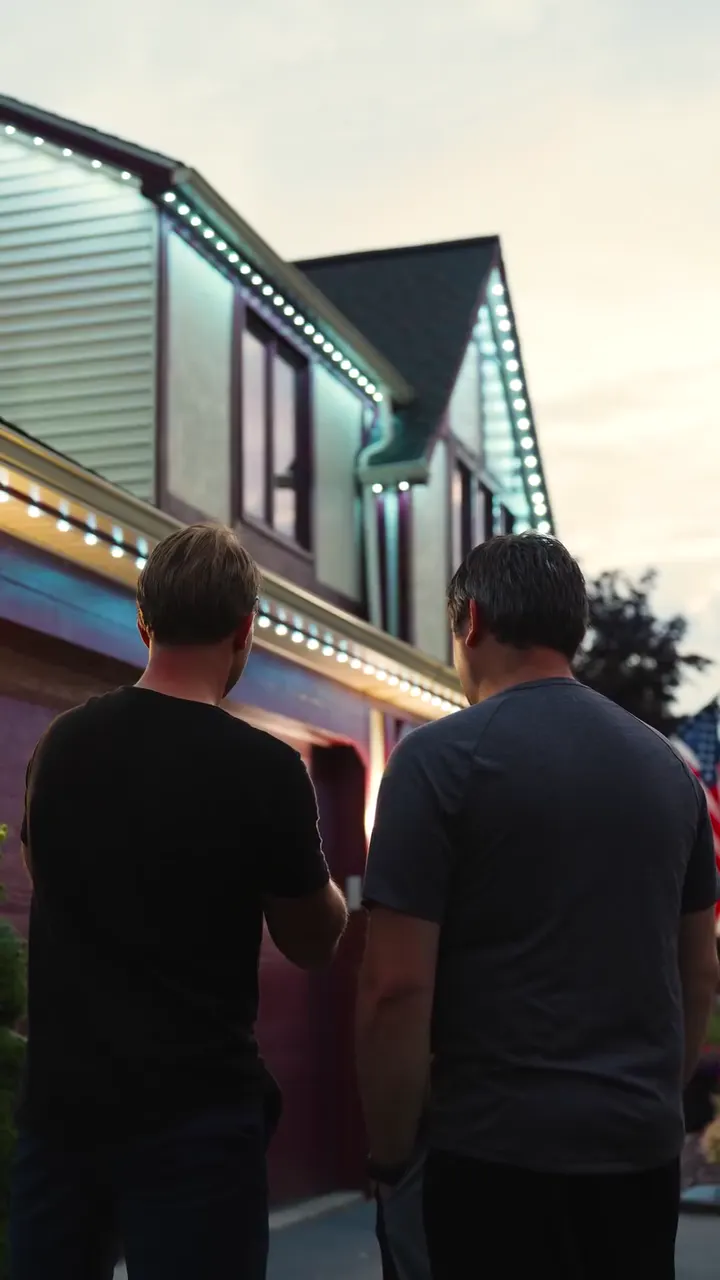 Two men in a driveway at dusk looking up at a lit roofline